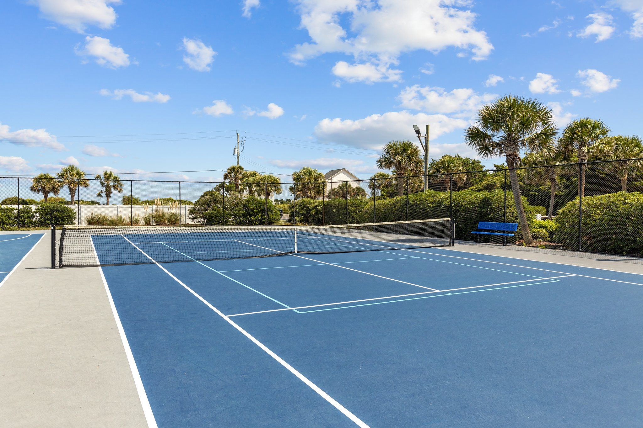 A tennis court with a net, surrounded by a fence, palm trees, and a bench.