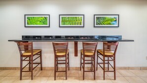 Une cuisine avec un plan de travail noir, trois chaises en bois dotées de coussins jaunes et trois tableaux représentant des feuilles vertes accrochés au mur.