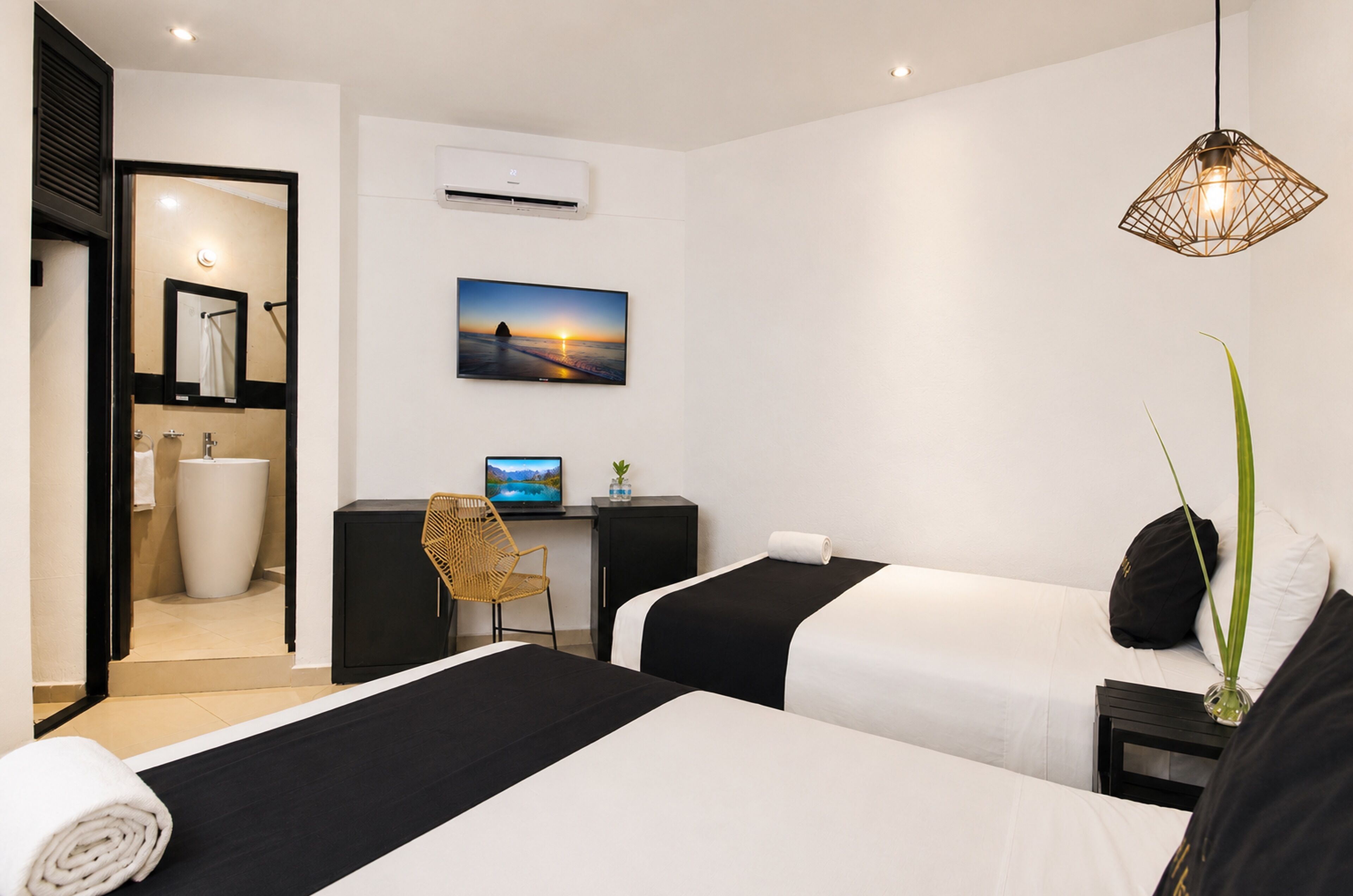Habitación de hotel con dos camas, un escritorio con computadora, baño y vista al atardecer.