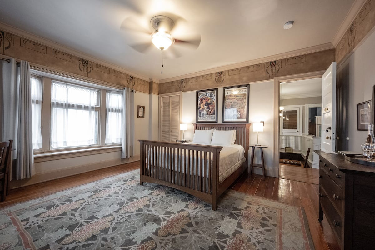 A bedroom with a wooden bed, a large window with curtains, a patterned rug, and a ceiling fan.
