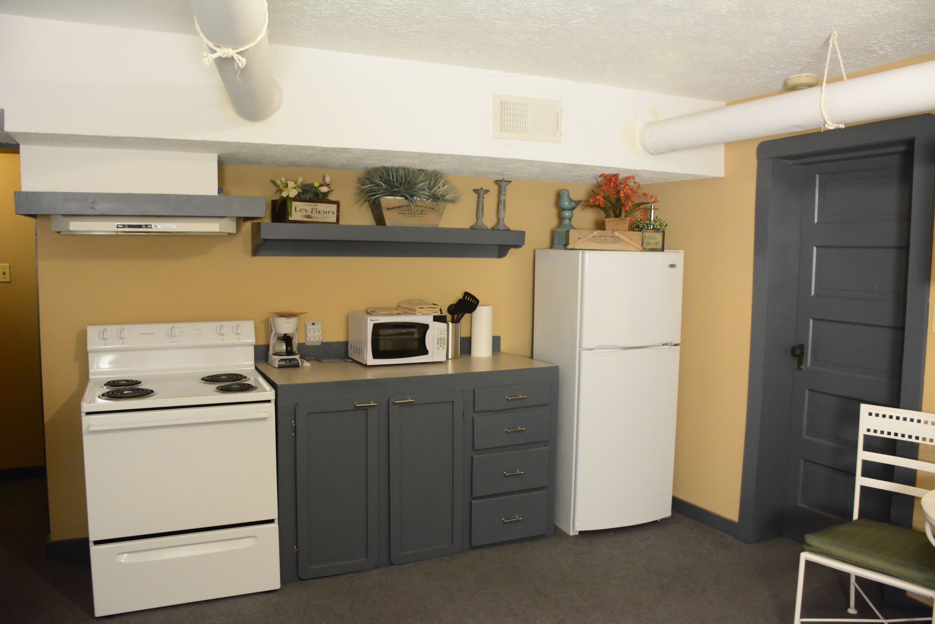 A compact kitchen with a stove, microwave, and refrigerator, featuring grey cabinets and decorative items on the countertop.