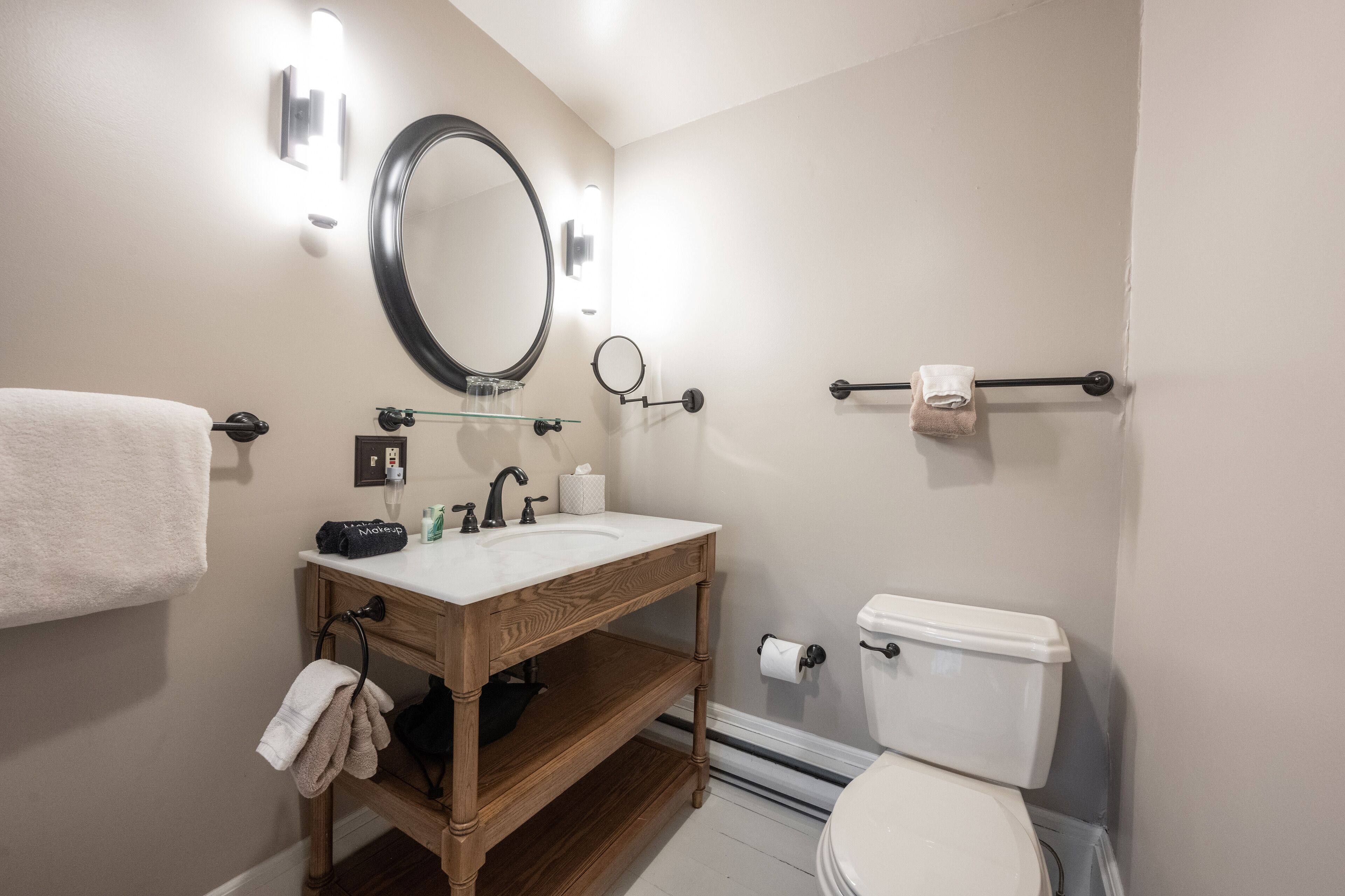 Un baño con tocador de madera, un espejo redondo, inodoro y toallero.