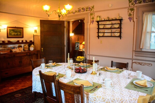 A dining room with a round table set for four, featuring a lace tablecloth, wooden chairs, and a fruit basket centerpiece.