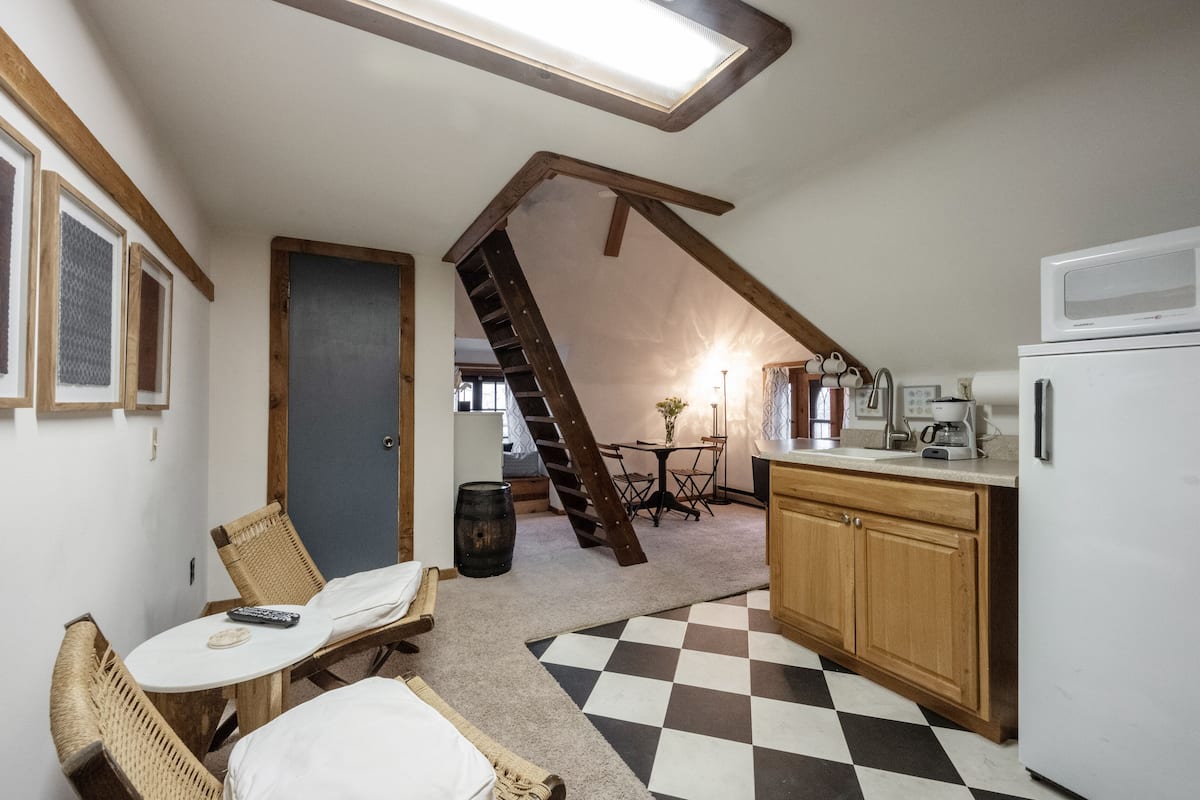 A compact kitchen with a refrigerator, microwave, and coffee maker. A wooden staircase leads to an upper level.