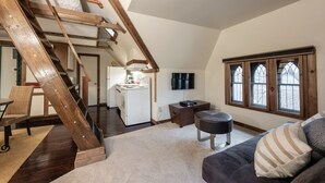 A loft-style room with a wooden staircase, a kitchenette, a sofa, and a window with stained glass.