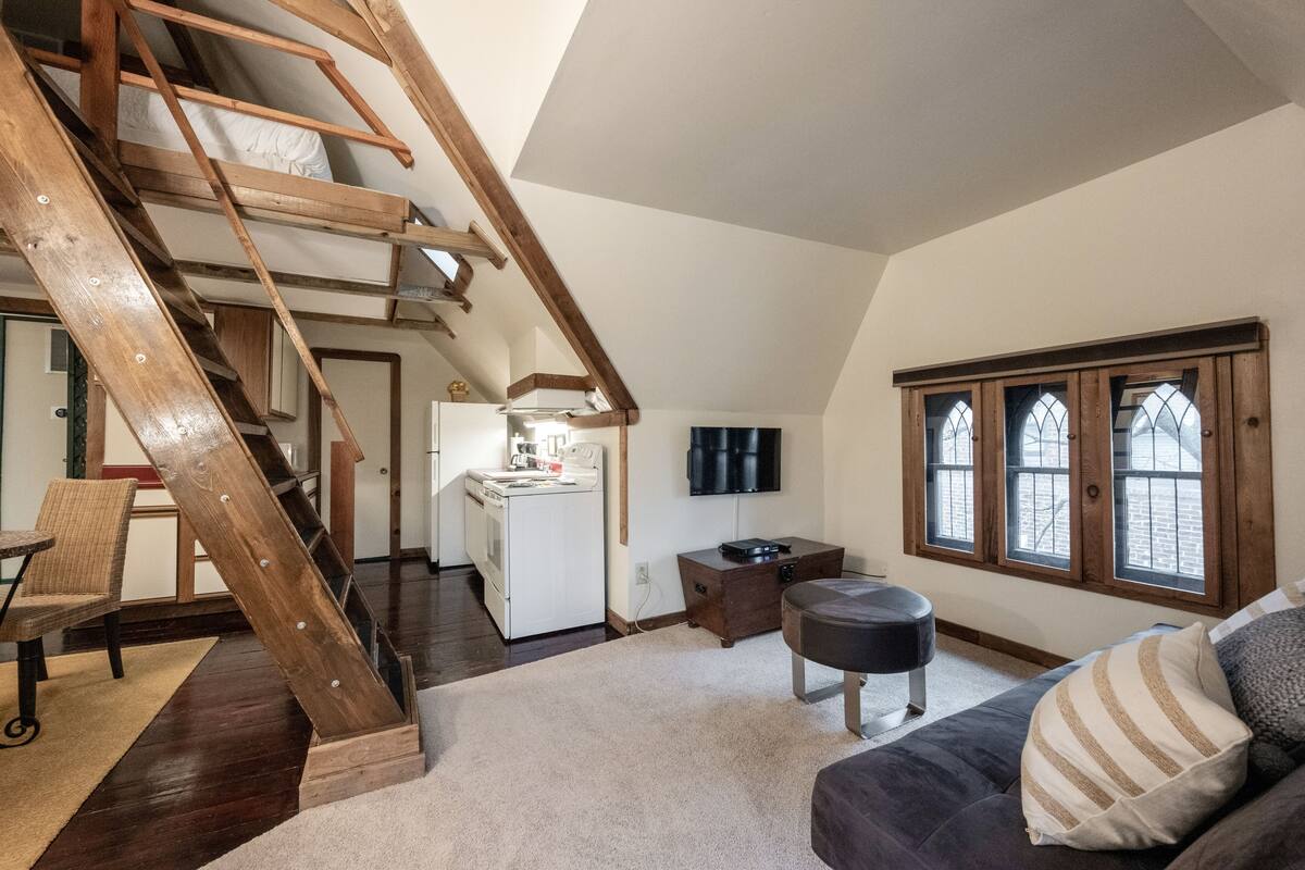 A loft-style room with a wooden staircase, a kitchenette, a sofa, and a window with stained glass.