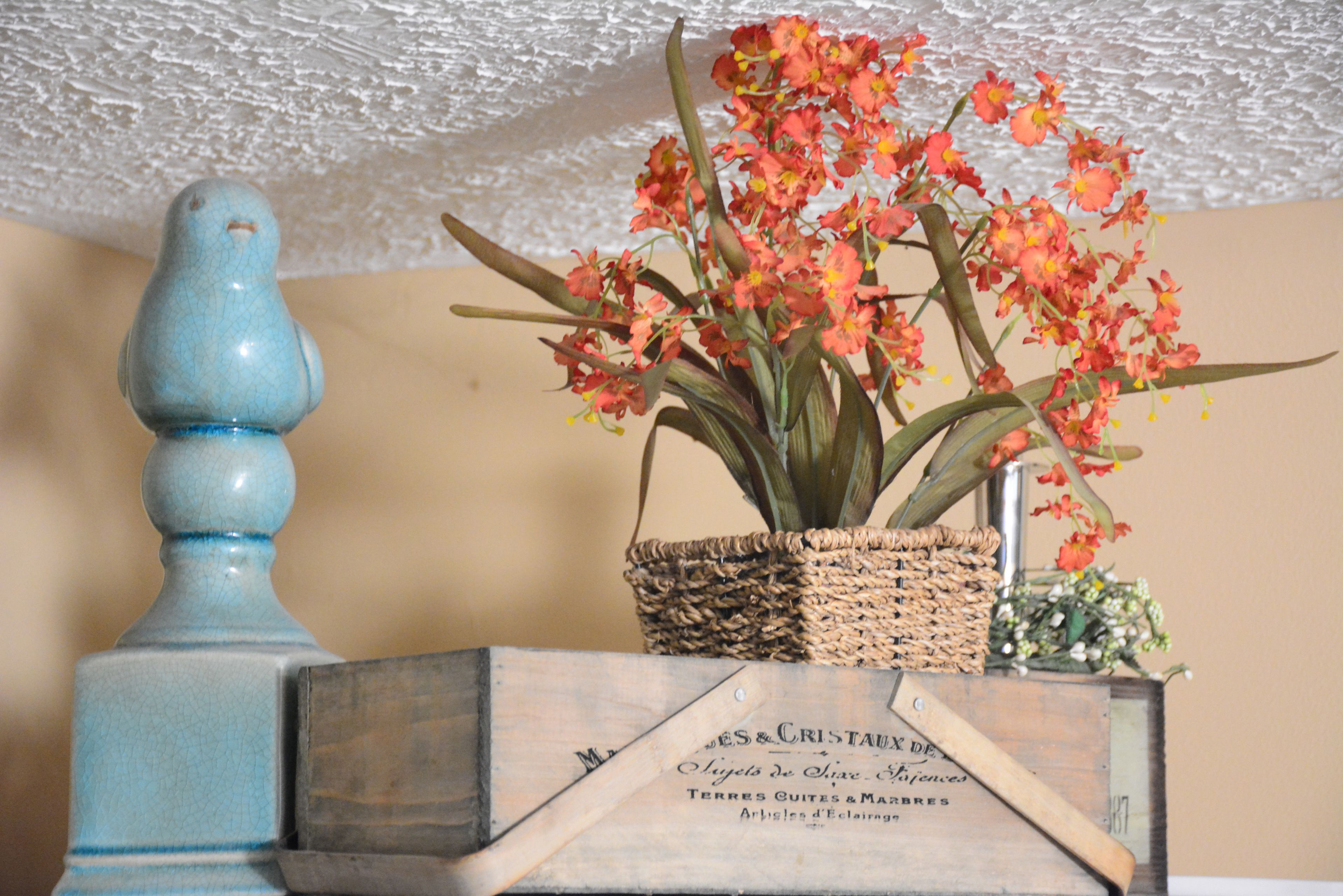 Un pájaro azul decorativo, una cesta de mimbre con flores rojas y una caja de madera con texto.