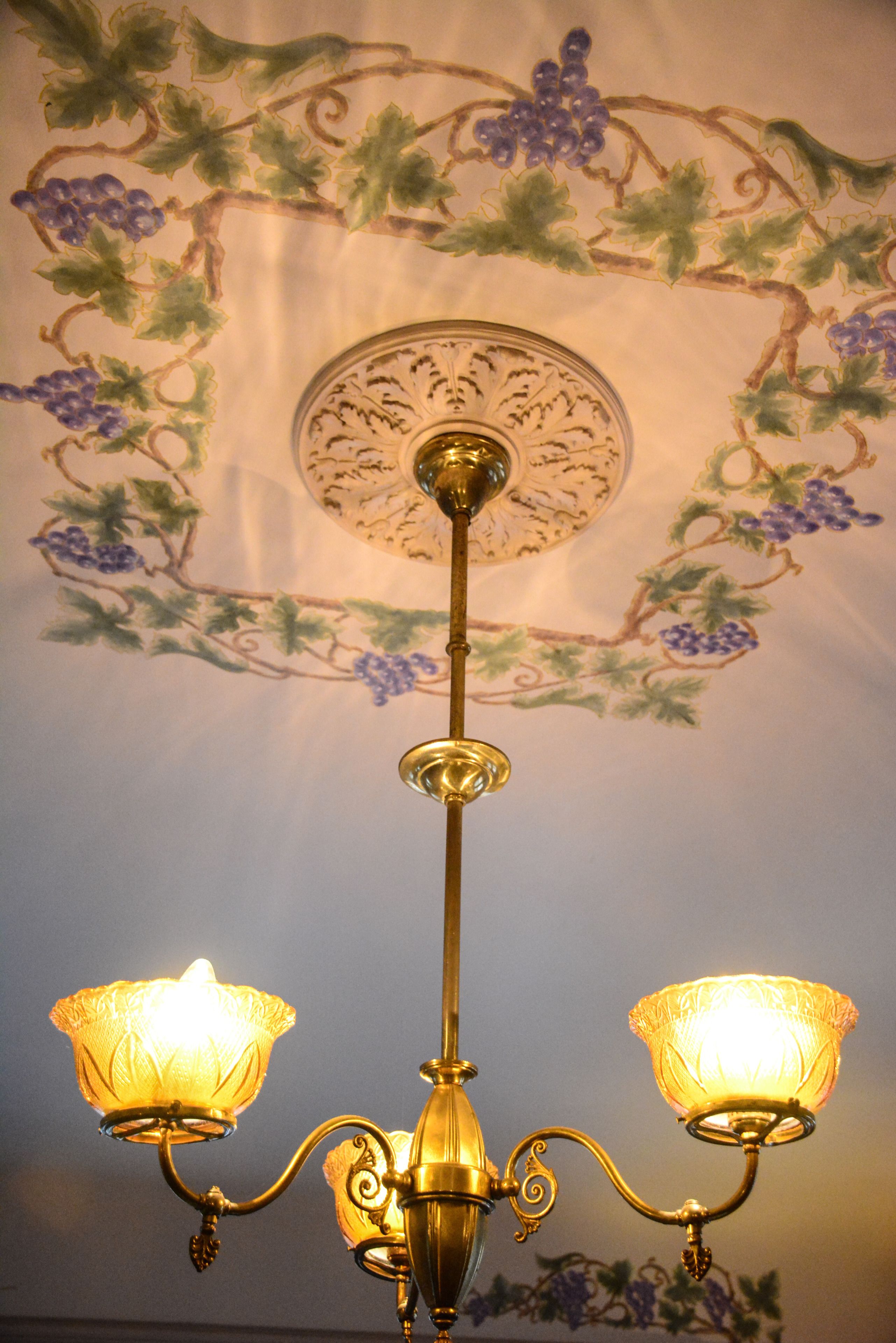 A chandelier with two illuminated lampshades and a wall mural with grapevines and grapes.