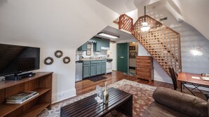 A spacious living area with a large flat-screen TV, wooden entertainment unit, and a cozy seating area. A staircase with a wooden railing leads to a kitchen area with teal cabinets and a dining table.