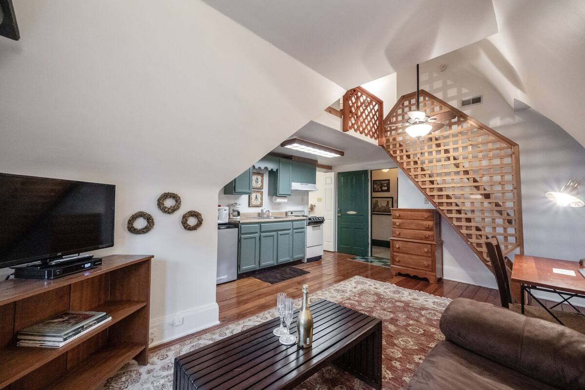 A spacious living area with a large flat-screen TV, wooden entertainment unit, and a cozy seating area. A staircase with a wooden railing leads to a kitchen area with teal cabinets and a dining table.