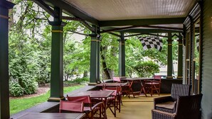 A covered patio area with wooden tables and chairs, green pillars, and a view of the surrounding greenery.