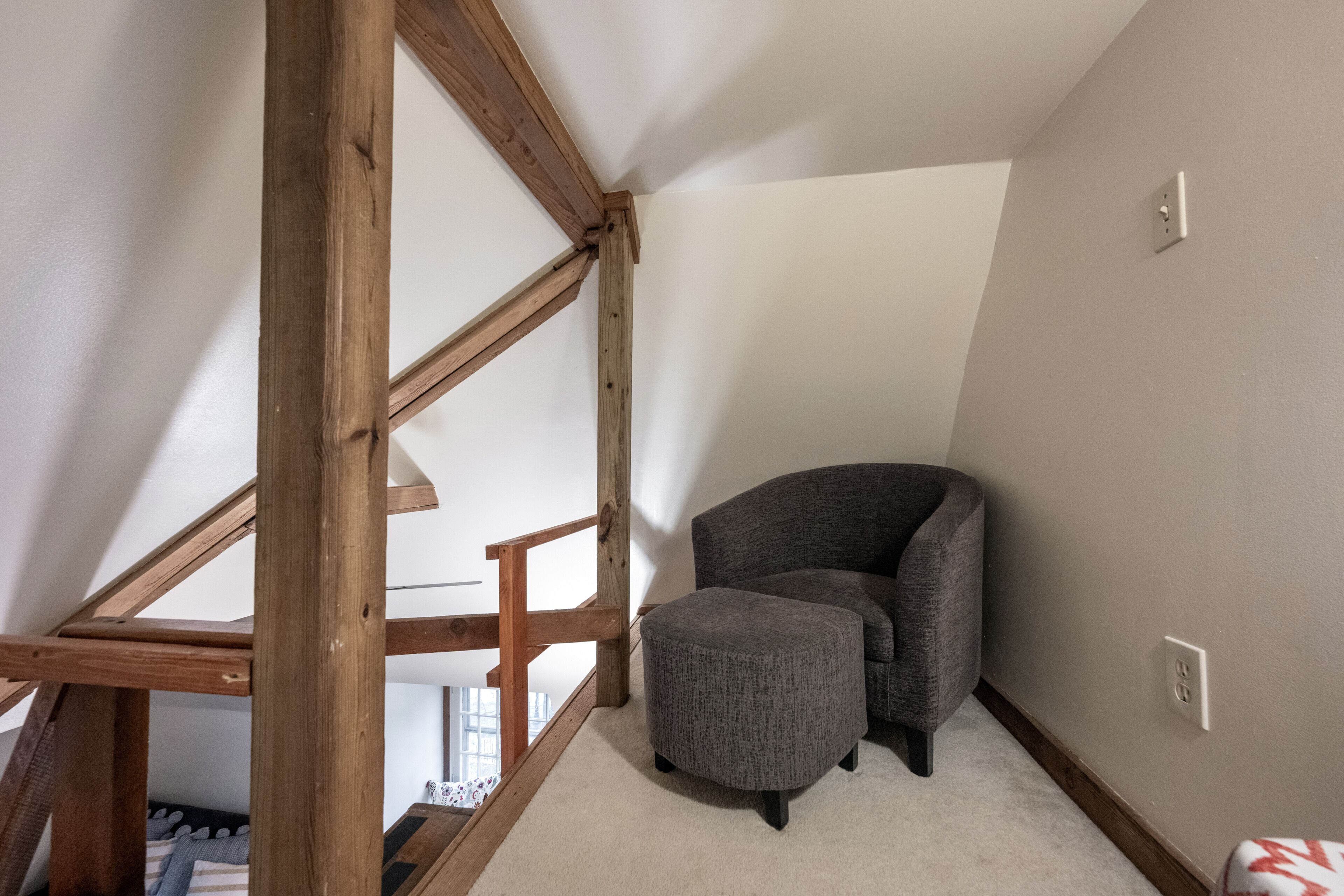 Une pièce d’angle avec un fauteuil et un pouf gris, des poutres en bois et un plafond blanc.