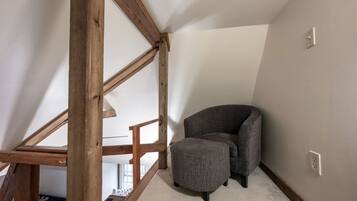 Une pièce d’angle avec un fauteuil et un pouf gris, des poutres en bois et un plafond blanc.