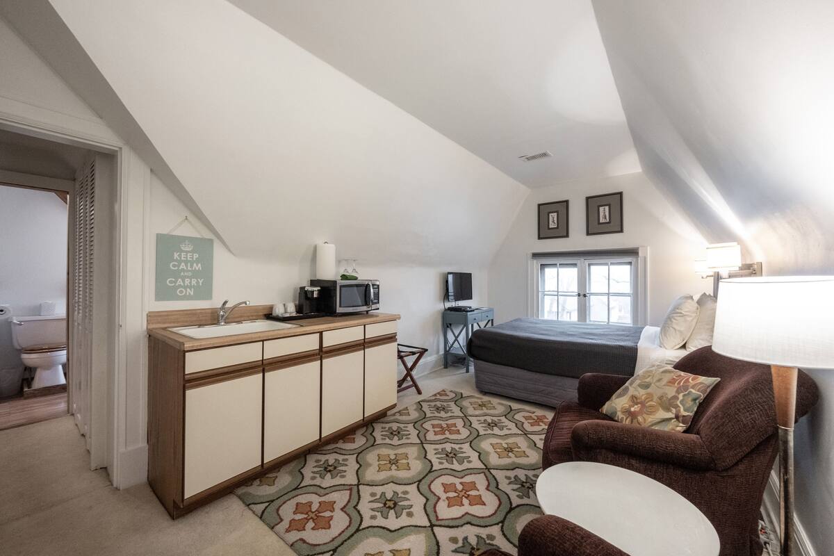 A bedroom with a bed, a desk with a computer, a chair, a window, and a door leading to the bathroom.