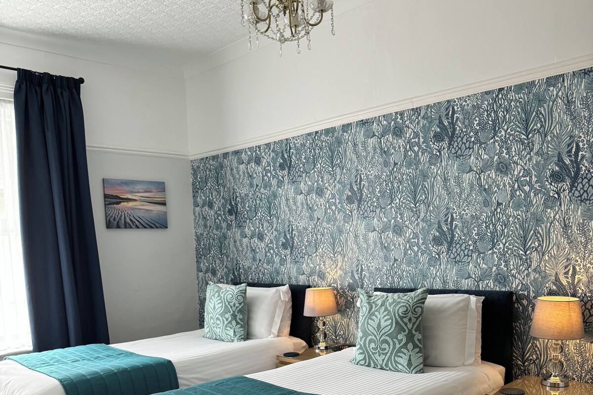 Ein Hotelzimmer mit zwei Betten, einem gemusterten Wandpapier, einer Kristallleuchtre und einem Nachttisch mit Lampe.
