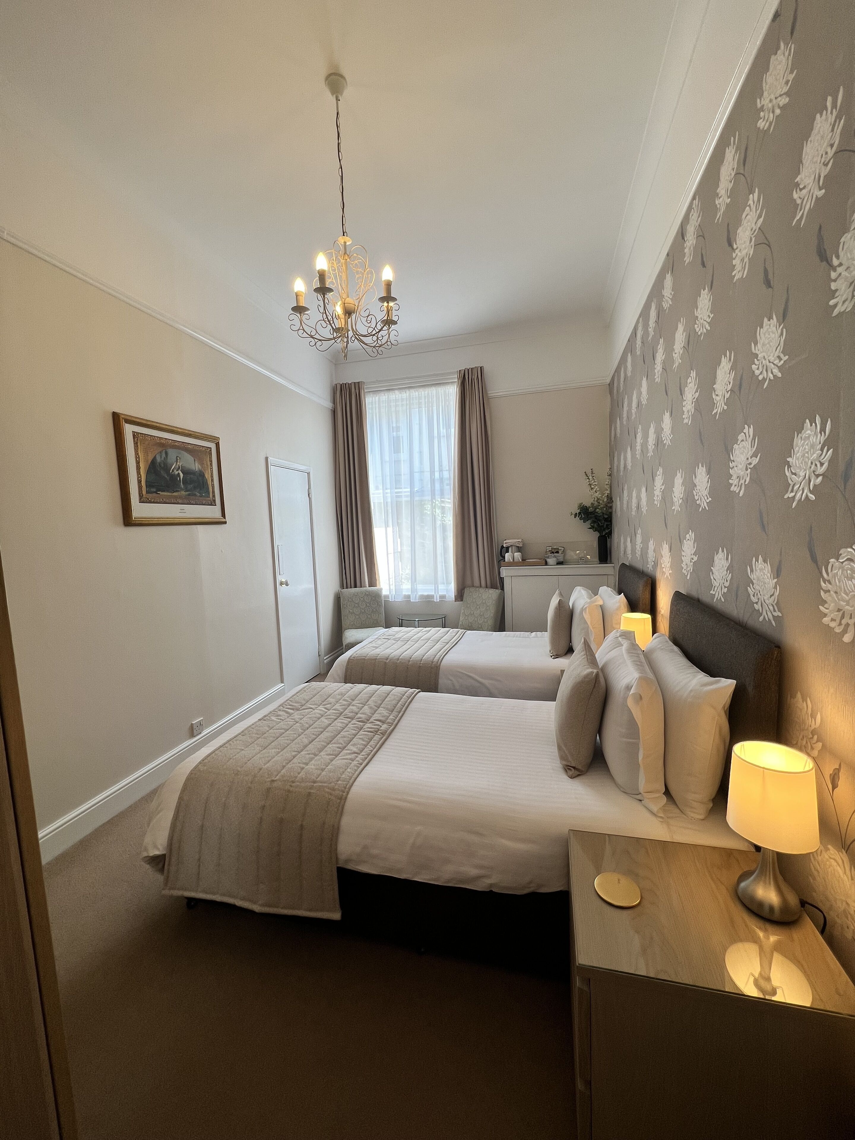 A hotel room with two beds, a chandelier, a bedside table with a lamp, a framed picture on the wall, and a window with curtains.