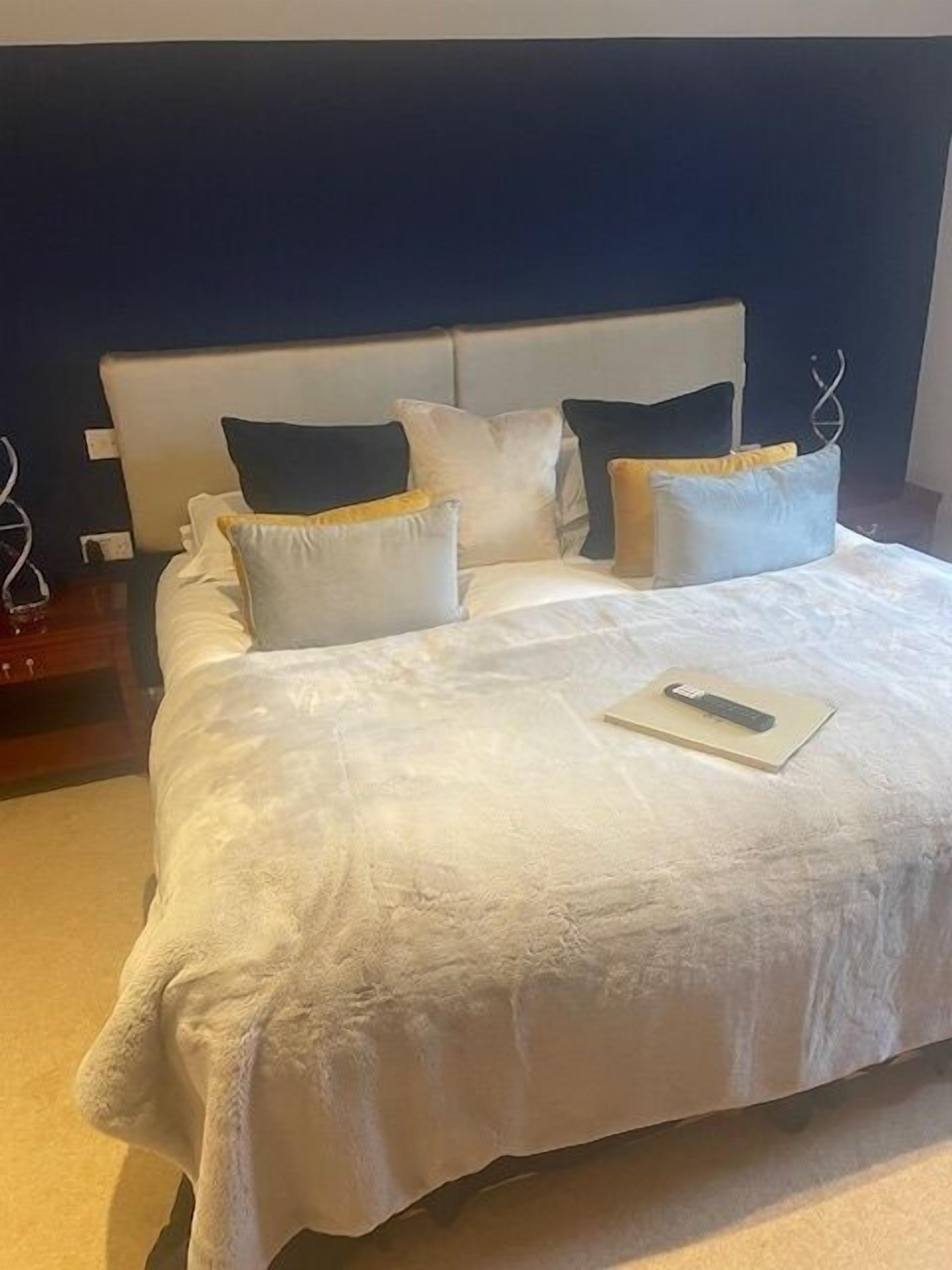 Una cama bien hecha con edredón blanco y varios almohadones, un control remoto sobre la cama y un cabecero de color oscuro.