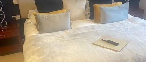 Una cama bien hecha con edredón blanco y varios almohadones, un control remoto sobre la cama y un cabecero de color oscuro.
