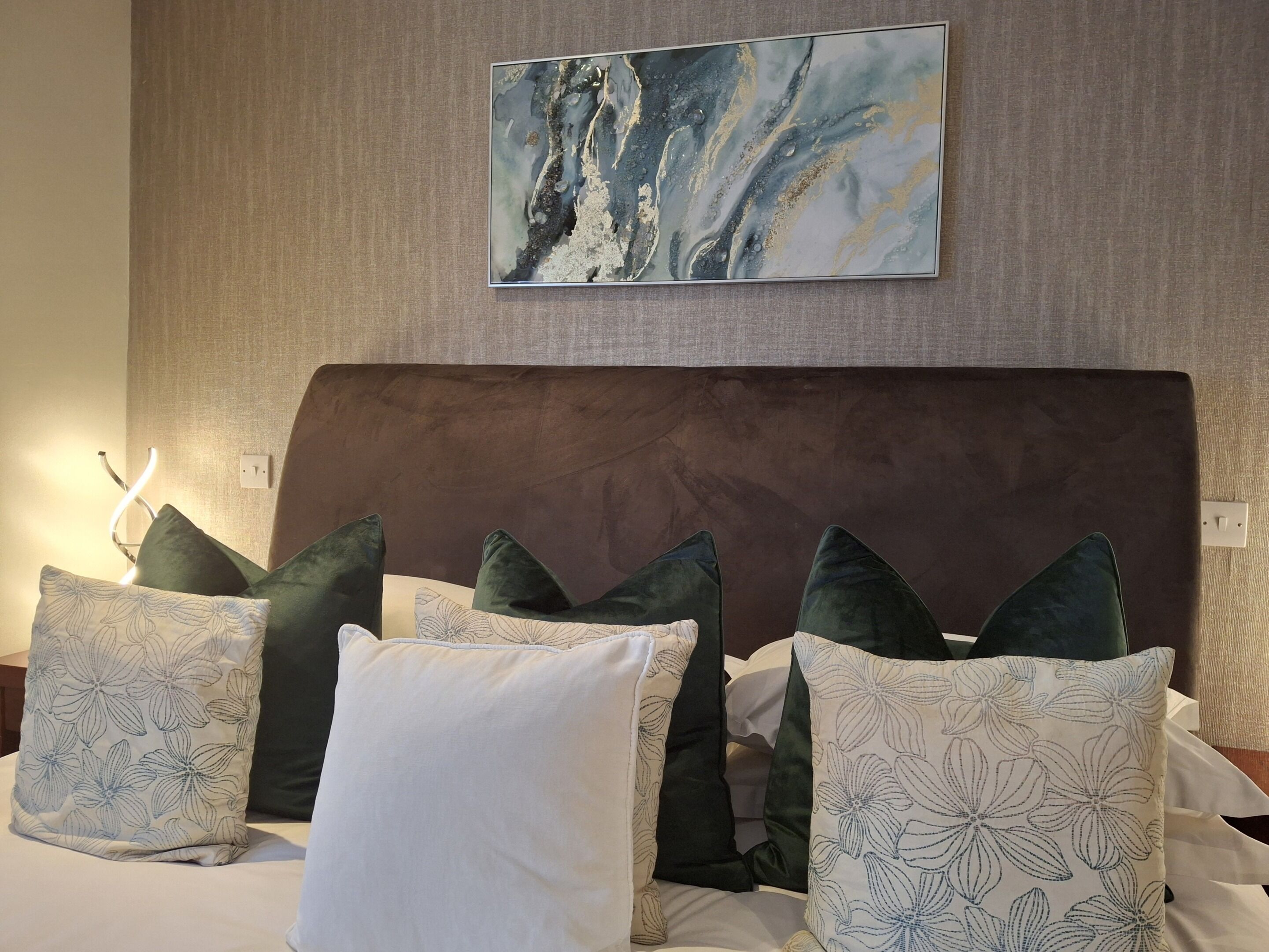 Una cama con cabecera marrón, varios almohadones y un cuadro abstracto colgado en la pared.