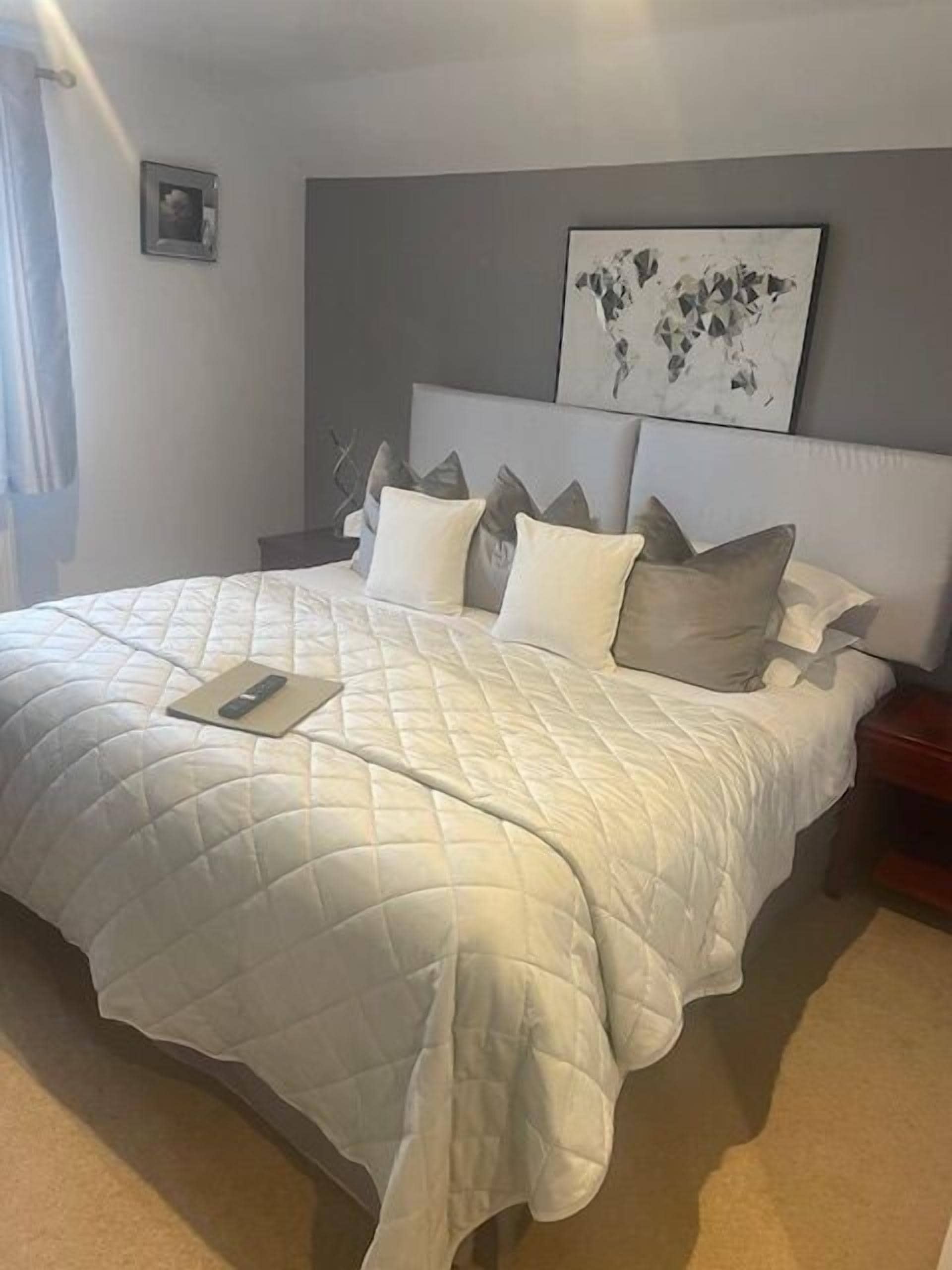 A neatly made bed with a quilted white duvet, a remote control, and several pillows. There is a framed picture on the wall above the bed.