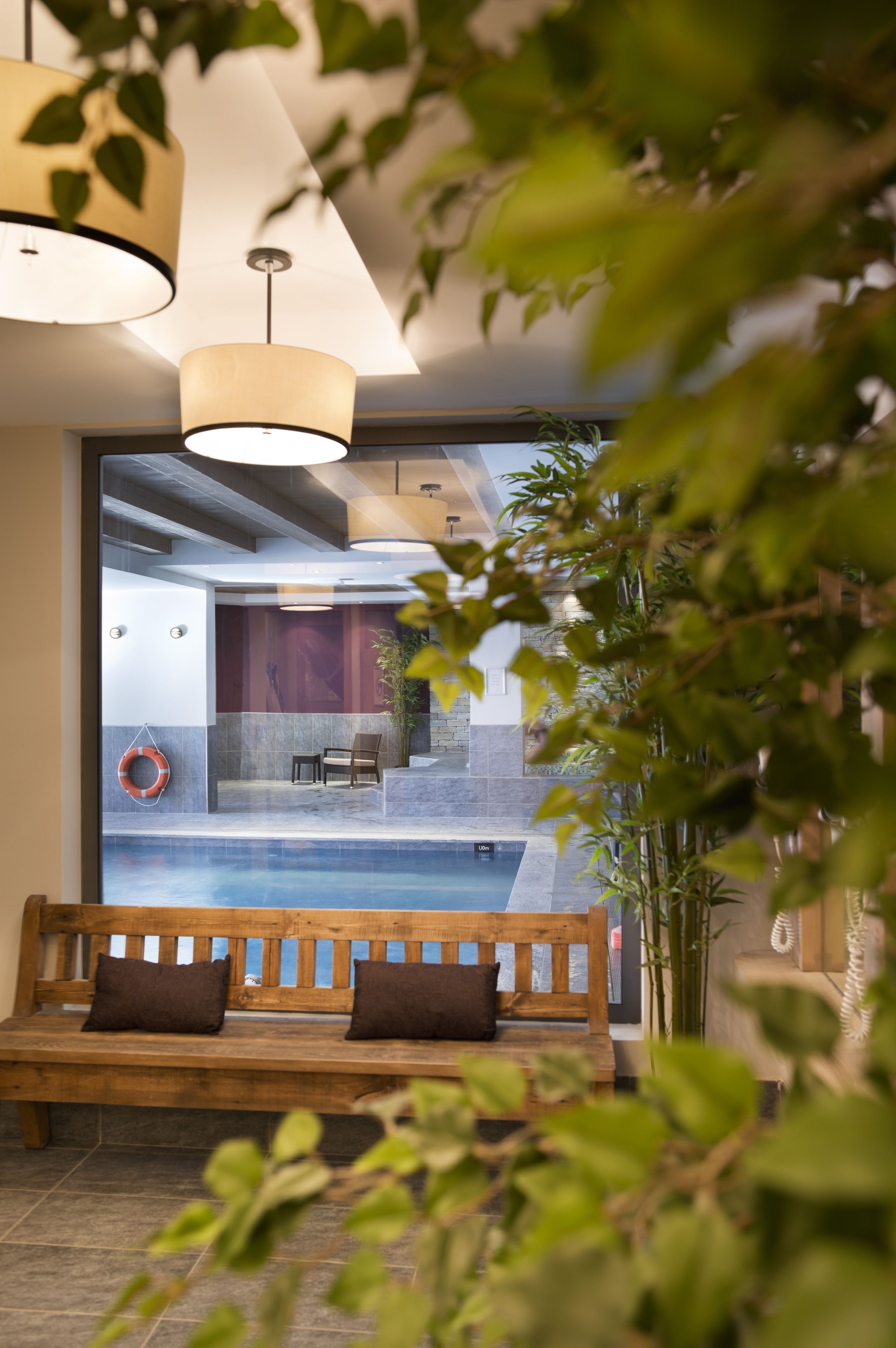 A modern interior with a wooden bench, a hanging life preserver, and a view of an indoor pool area.