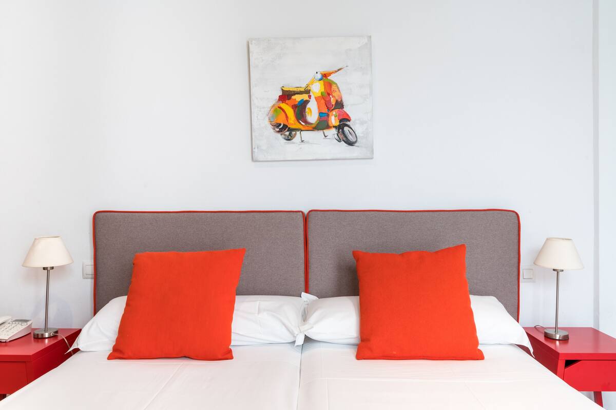 Una cama doble con almohadas blancas y rojas, un cabecero gris y una mesita de noche roja con una lámpara.