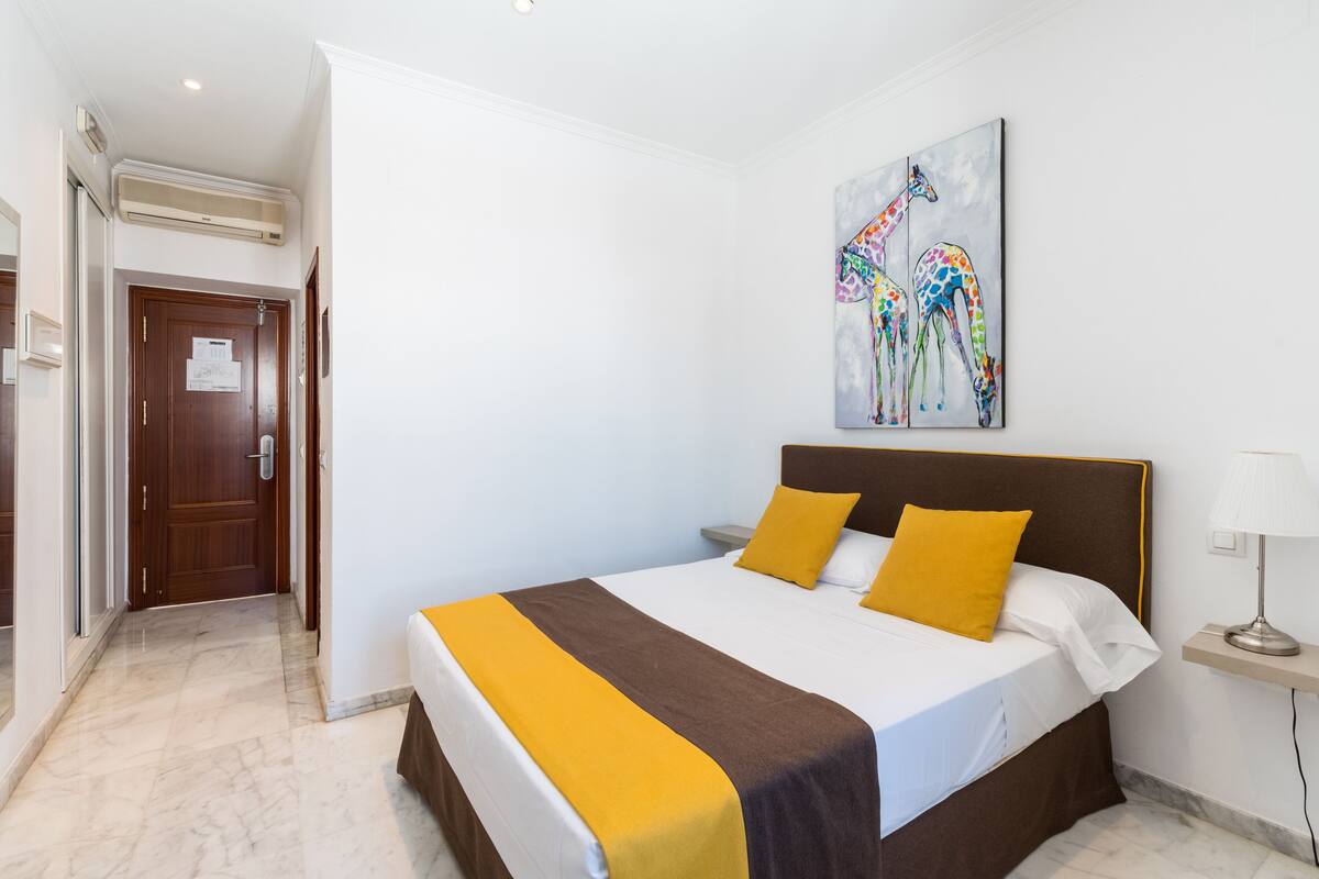 Habitación de hotel con cama, mesita de noche, lámpara y un cuadro de jirafas en la pared.