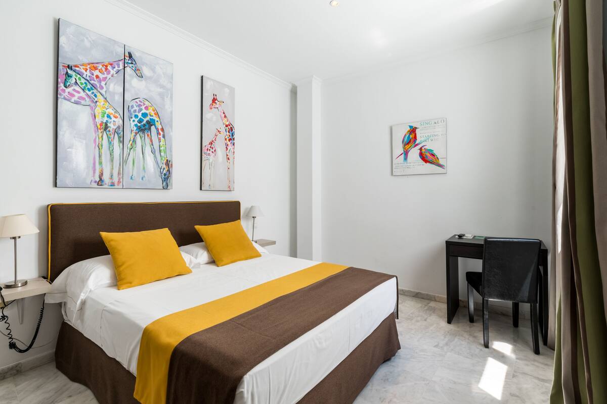Habitación de hotel con una cama grande, dos almohadas amarillas, un escritorio con silla y obras de arte en las paredes.