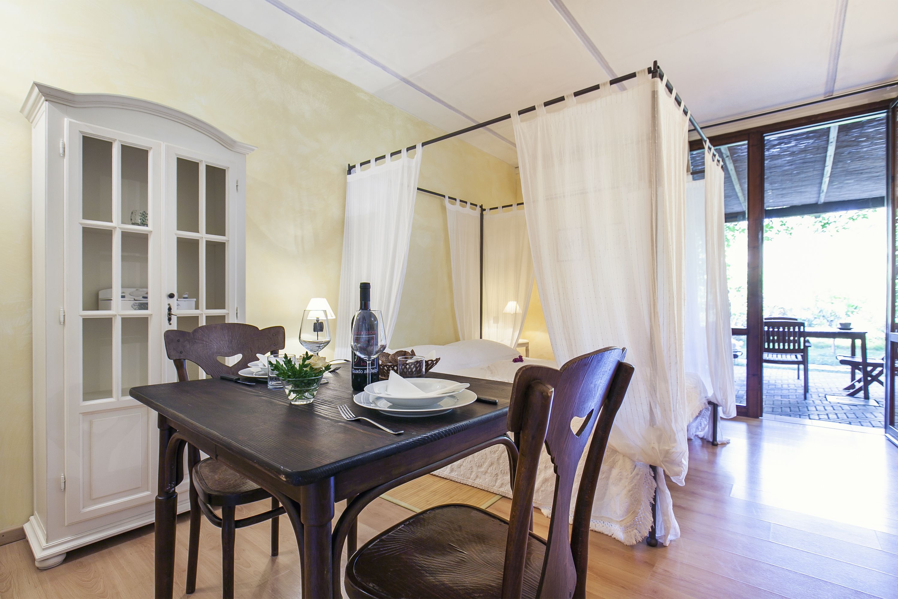 Un comedor con una mesa preparada para dos, un aparador con puertas de vidrio y una cama con dosel y cortinas blancas.