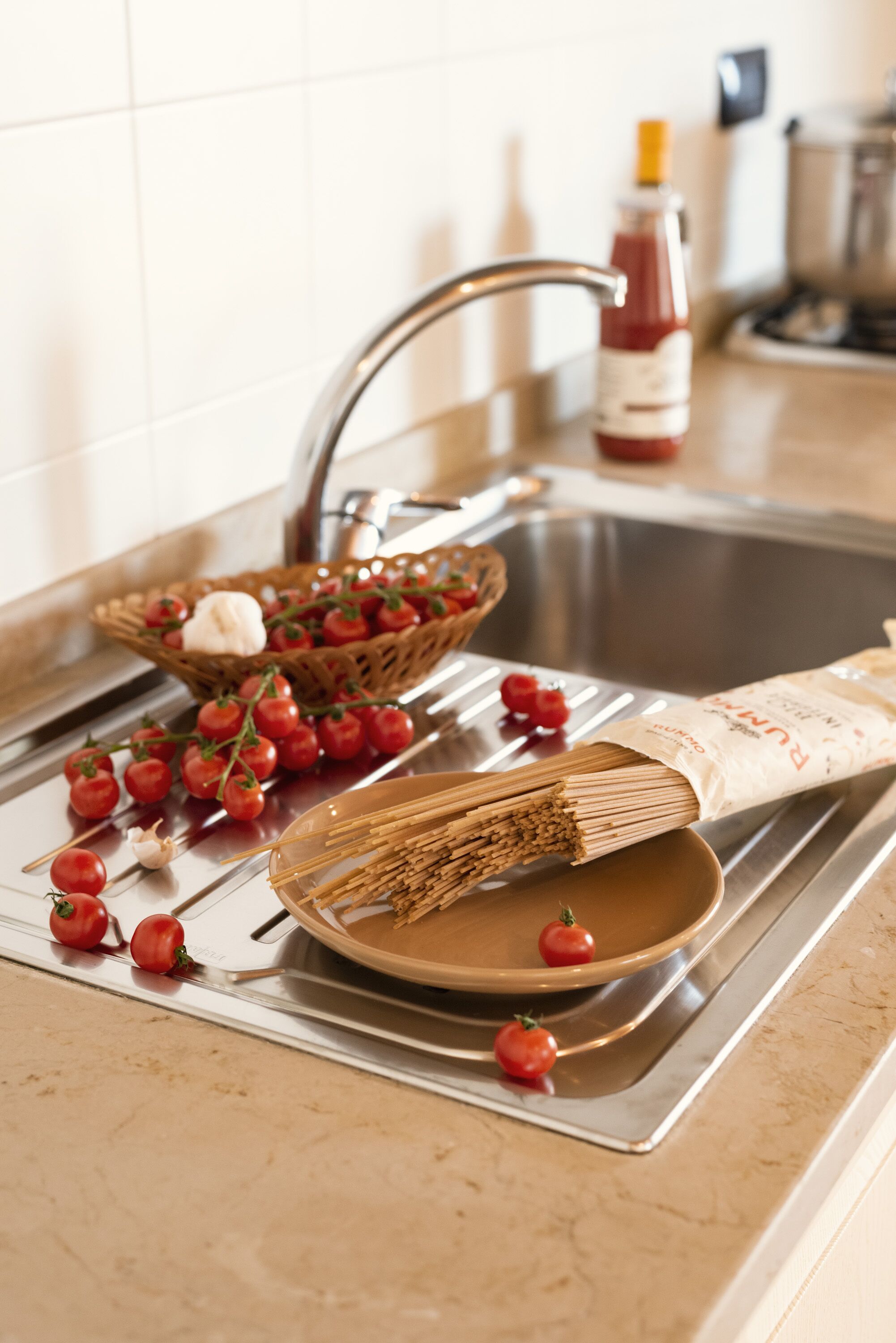 A kitchen sink with a basket of tomatoes, a bowl of pasta, and a bottle of ketchup.