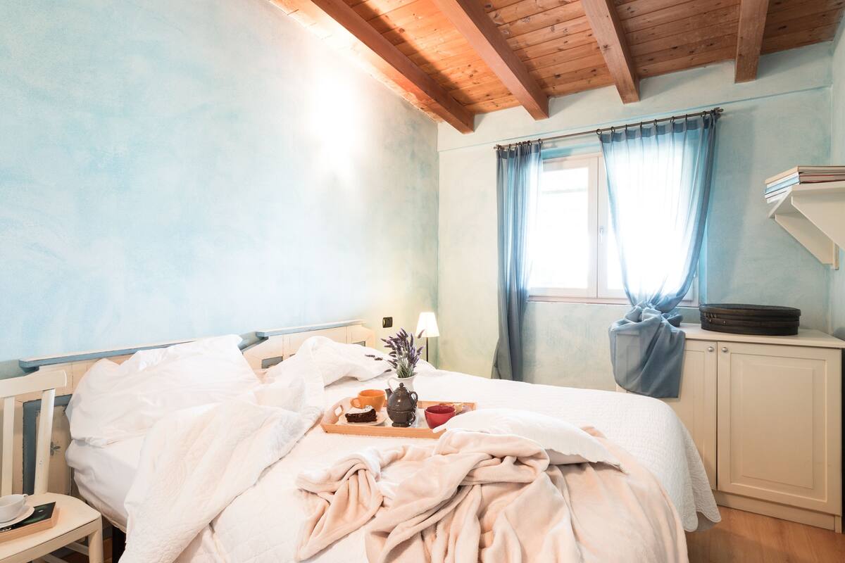 Quarto com teto de madeira, cama com roupa de cama branca, bandeja com itens de café da manhã, mesa de cabeceira e janela com cortinas.