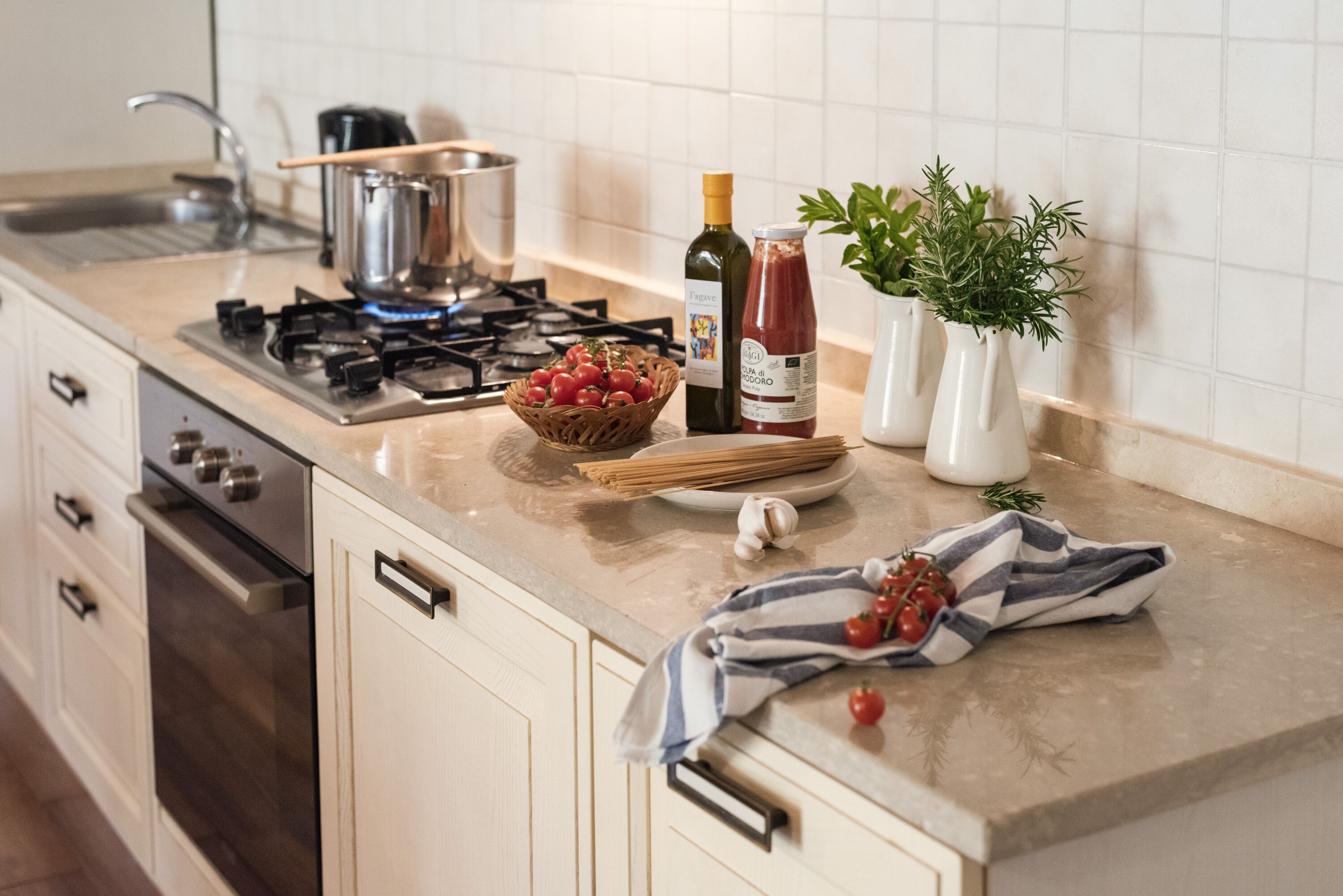 Une cuisine équipée d’une cuisinière à gaz, d’un évier, d’un panier de tomates, d’une bouteille d’huile d’olive, d’une bouteille de sauce tomate et d’une plante en pot.