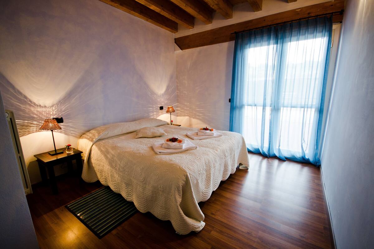 Quarto com uma cama grande, duas mesas de cabeceira, um criado-mudo, uma janela com cortinas leves e teto de madeira.