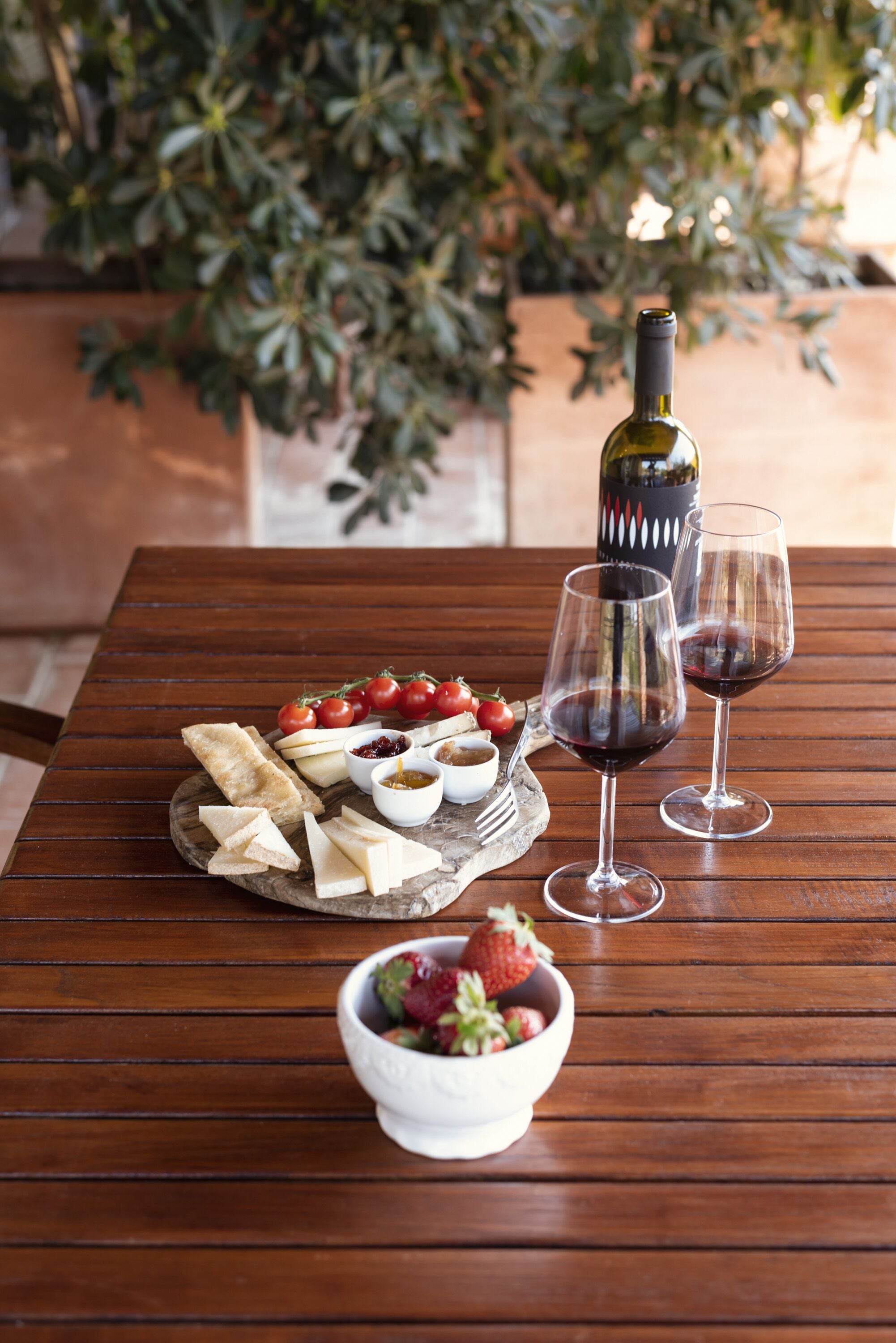 Une table en bois avec une bouteille de vin, deux verres à vin, une assiette de fromage et un bol de fraises.