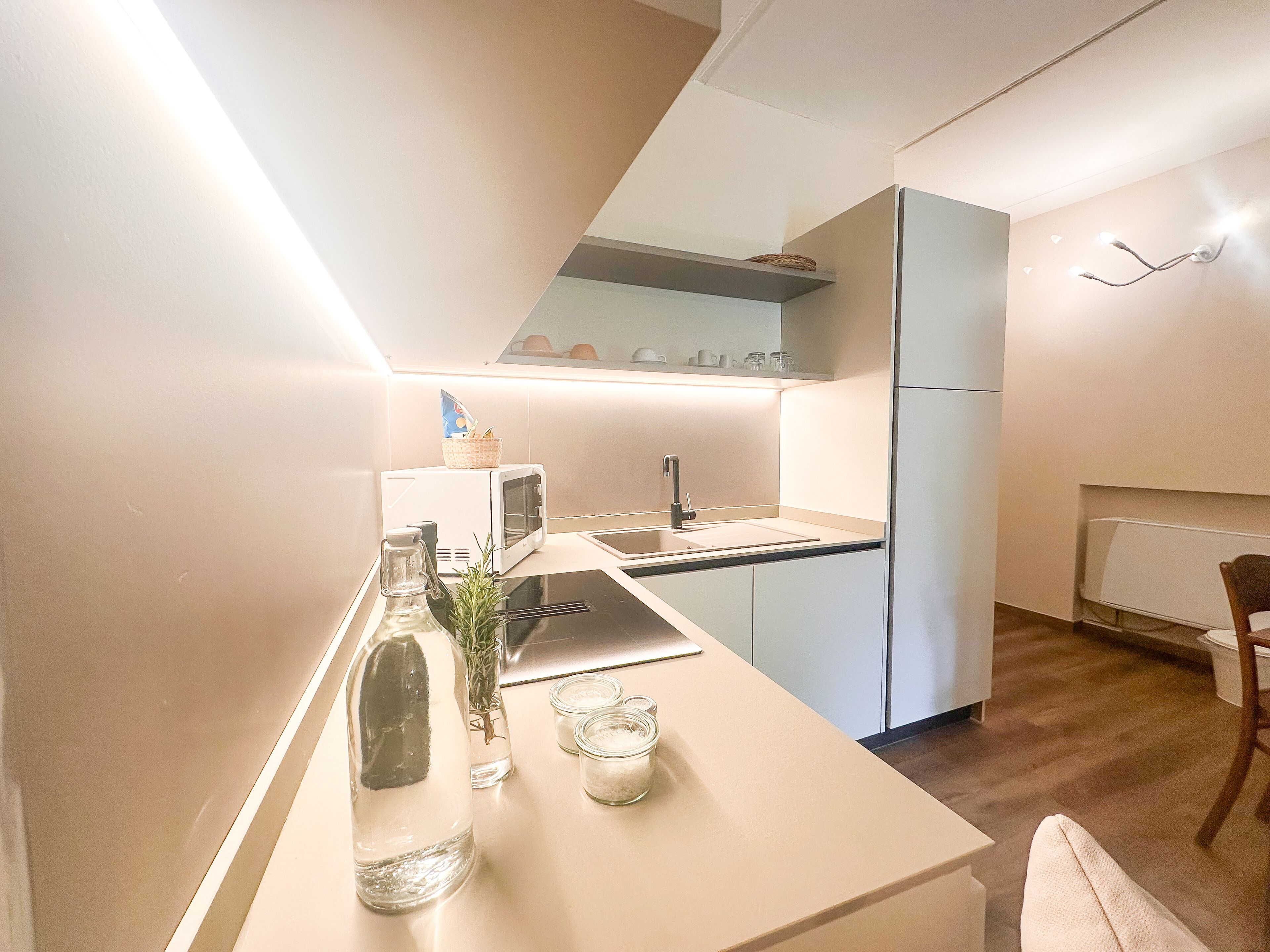 Une cuisine moderne avec un plan de travail blanc, un four à micro-ondes encastré et un évier. On peut voir une bouteille d’eau et un verre posés sur le plan de travail.