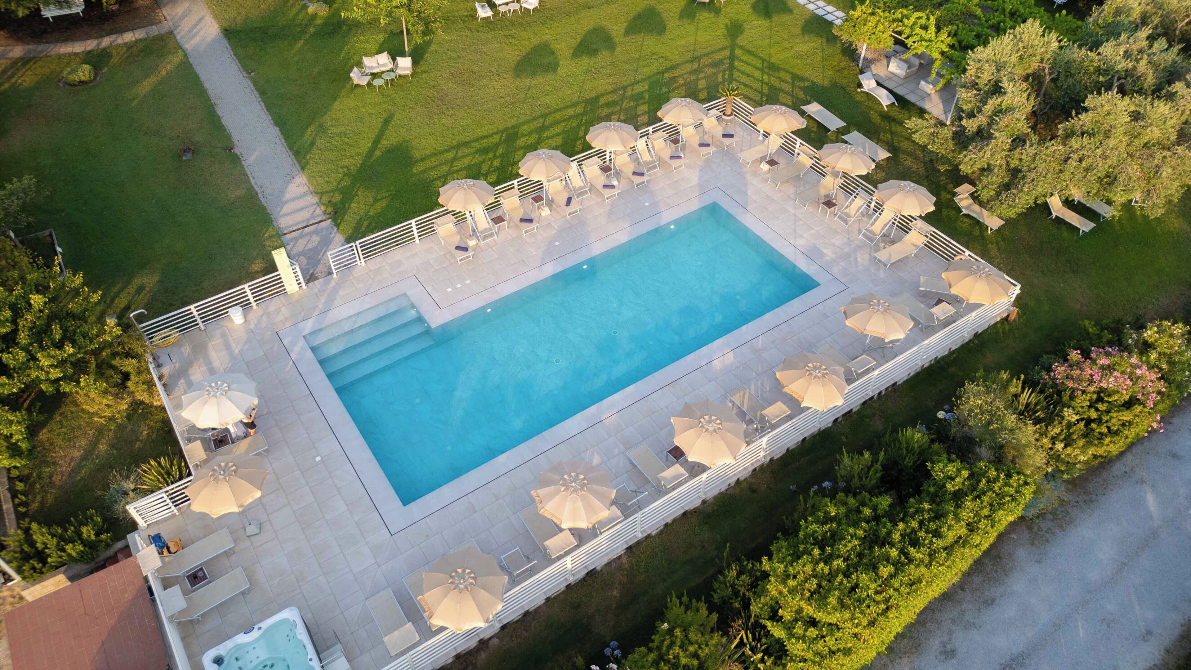 An aerial view of a rectangular swimming pool with a tiled deck, surrounded by lounge chairs and umbrellas, and bordered by a well-maintained lawn.