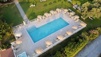 An aerial view of a rectangular swimming pool with a tiled deck, surrounded by lounge chairs and umbrellas, and bordered by a well-maintained lawn.