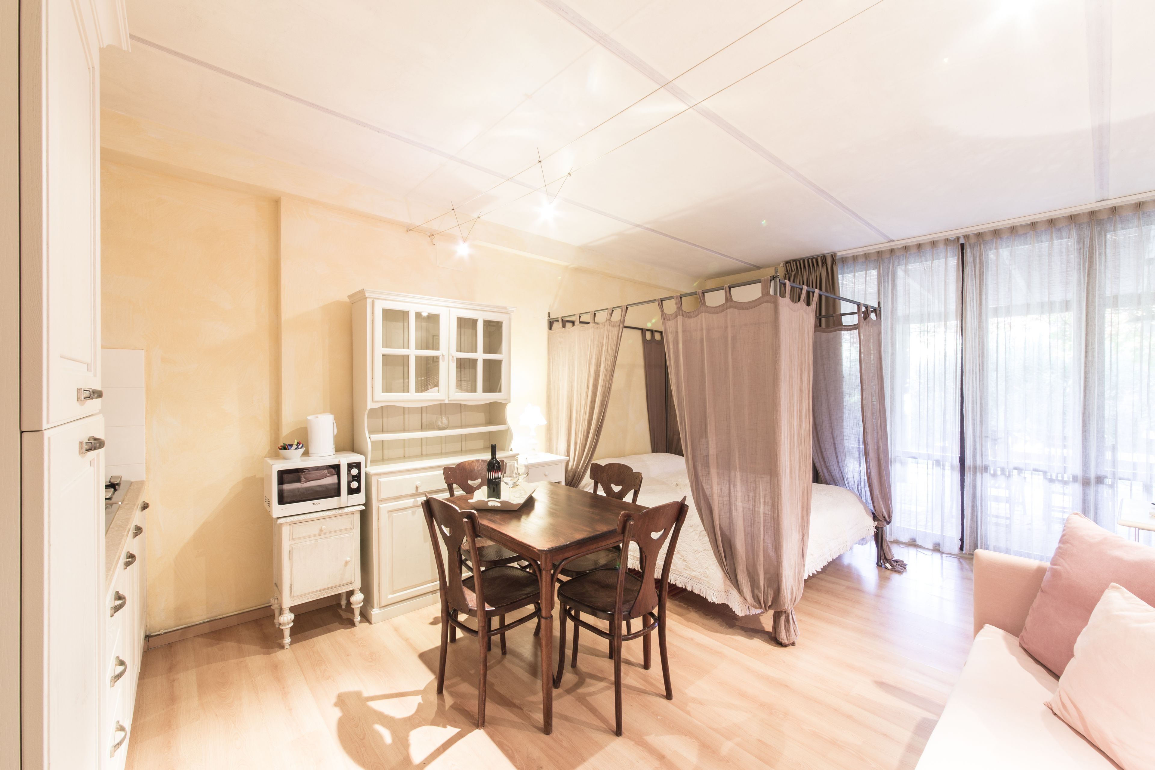 A bedroom with a bed, a bedside table, a window with curtains, a small table with chairs, and a microwave.