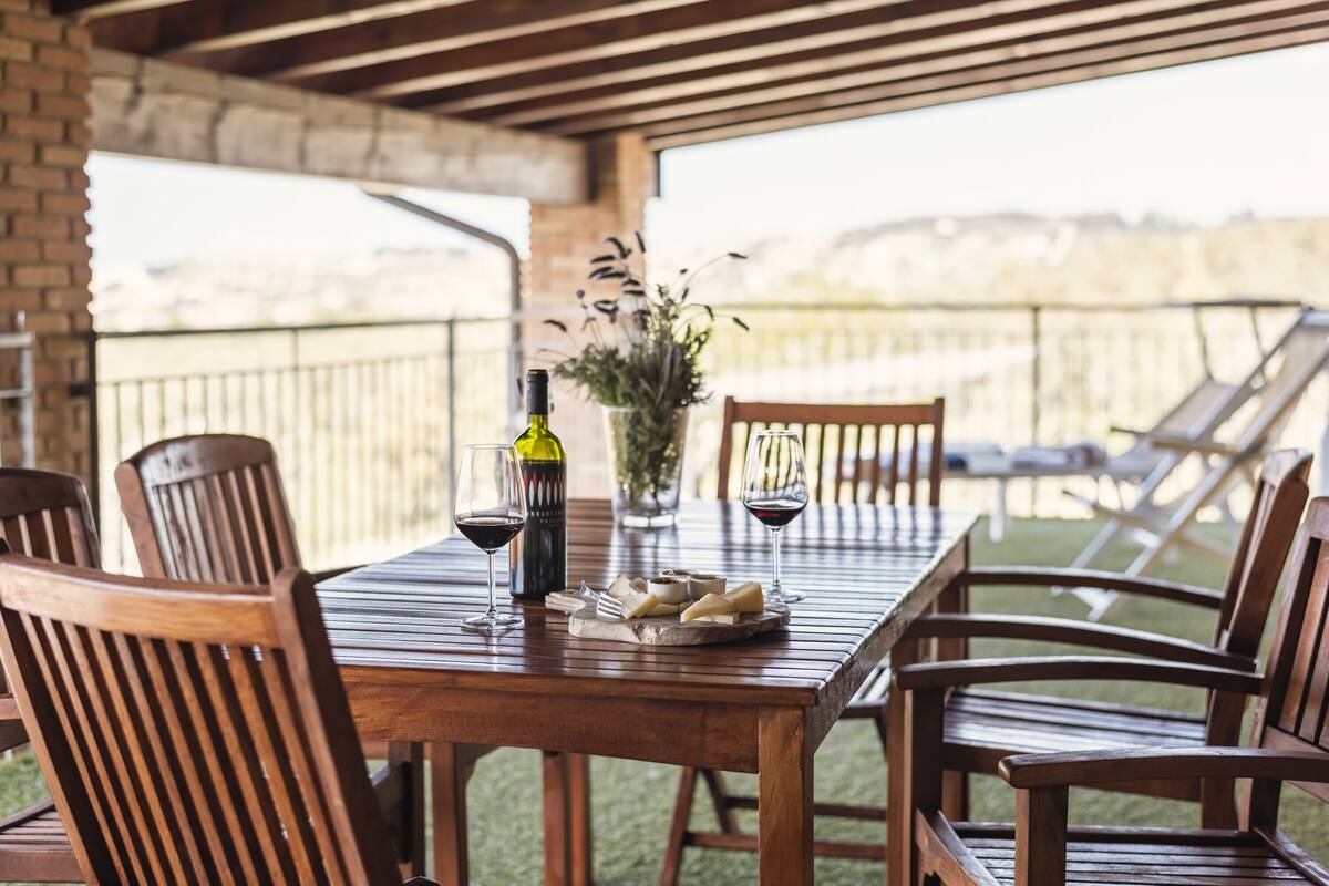 Uma mesa de madeira com cadeiras, uma garrafa de vinho e copos em um pátio com vista.