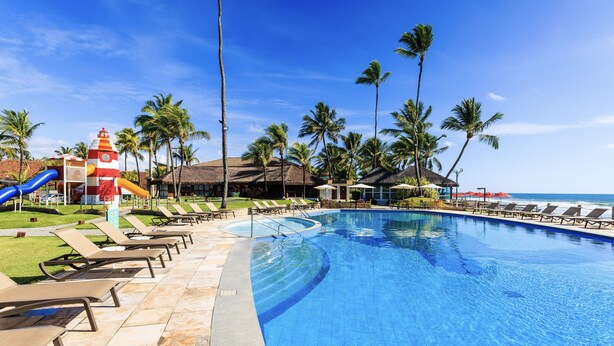 A swimming pool with lounge chairs and palm trees, a slide, and a thatched-roof building. - Armação Resort Porto de Galinhas (Ipojuca)