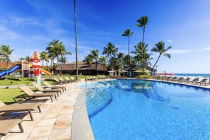 A swimming pool with lounge chairs and palm trees, a slide, and a thatched-roof building. - Armação Resort Porto de Galinhas (Ipojuca)