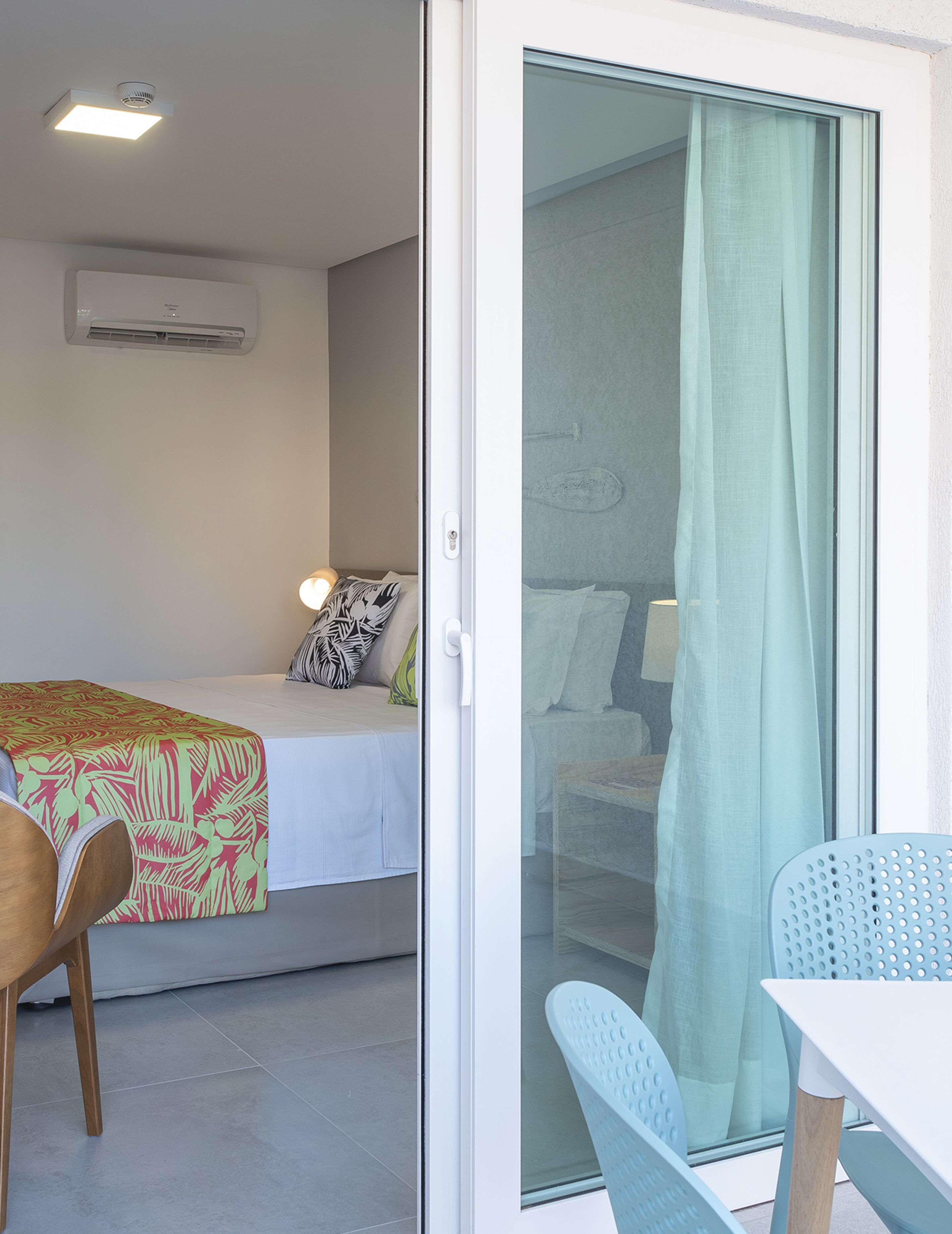 A room with a bed, a chair, a table, and a sliding glass door leading to an outdoor area.