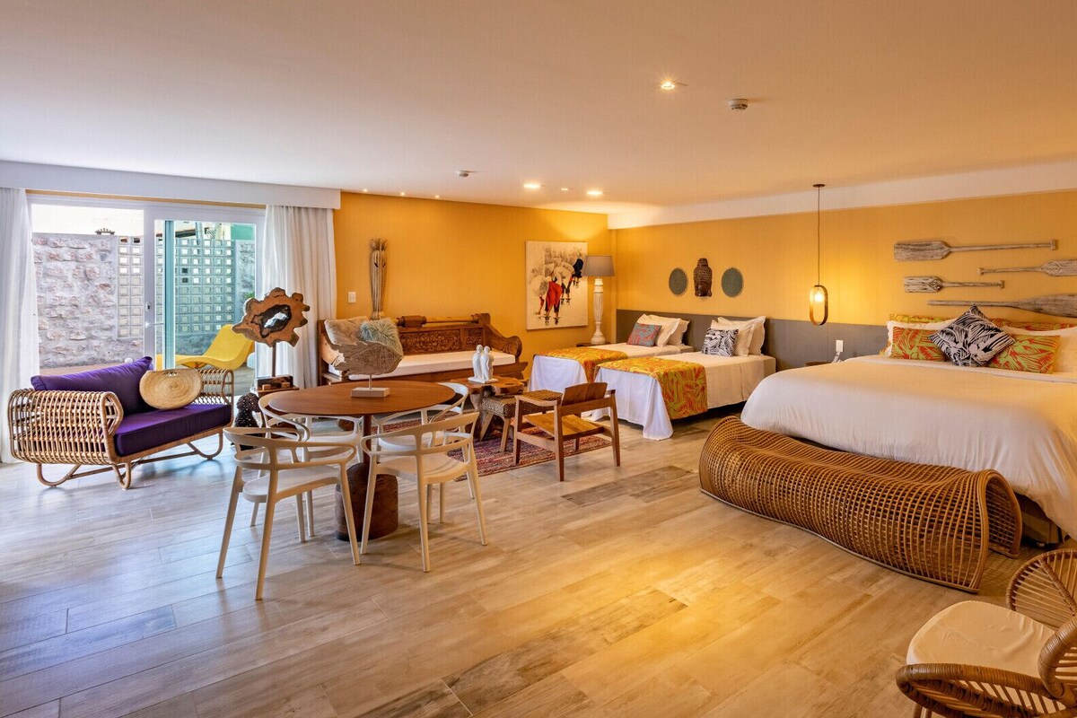 A hotel room with a bed, two bedside tables, a sofa, a dining table with chairs, and a balcony with a chair.