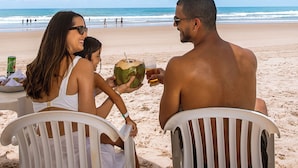 A beach scene with a white umbrella, two people sitting on white chairs, and a man holding a coconut drink. - Armação Resort Porto de Galinhas (Ipojuca)