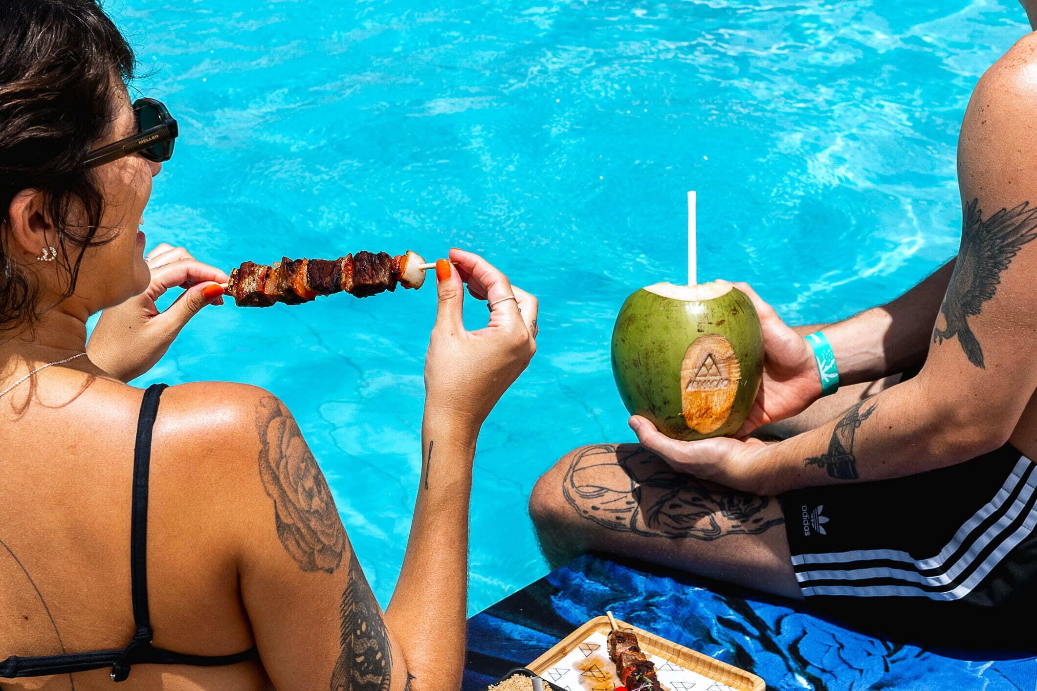 Dos personas junto a una piscina, una sosteniendo un pincho de comida y la otra, un coco con una pajita.