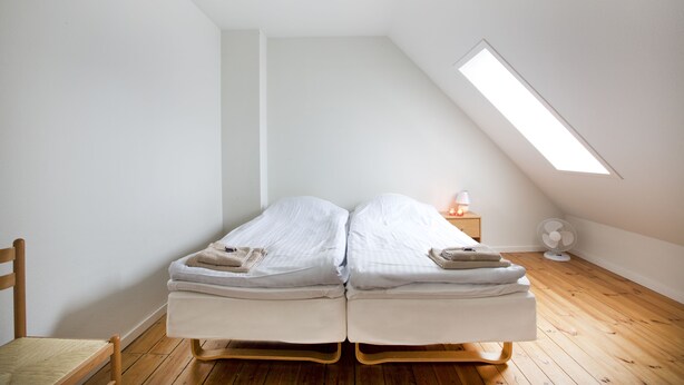 A small, single-room apartment with a sloped ceiling, two single beds, a wooden floor, a chair, a bedside table with a lamp, and a wall-mounted fan. - Motel Stege (Stege)