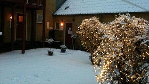 A snow-covered courtyard with a wooden balcony and a snow-covered bush with lights. - Motel Stege (Stege)