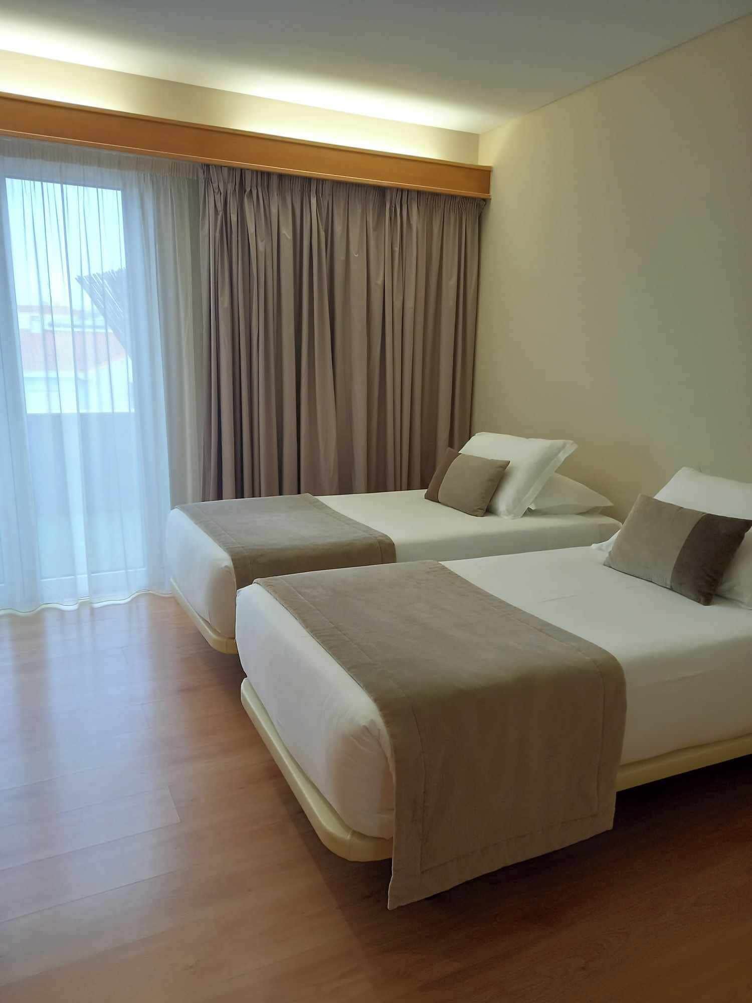 Habitación de hotel con dos camas, un ventanal grande con cortinas y suelo de madera.