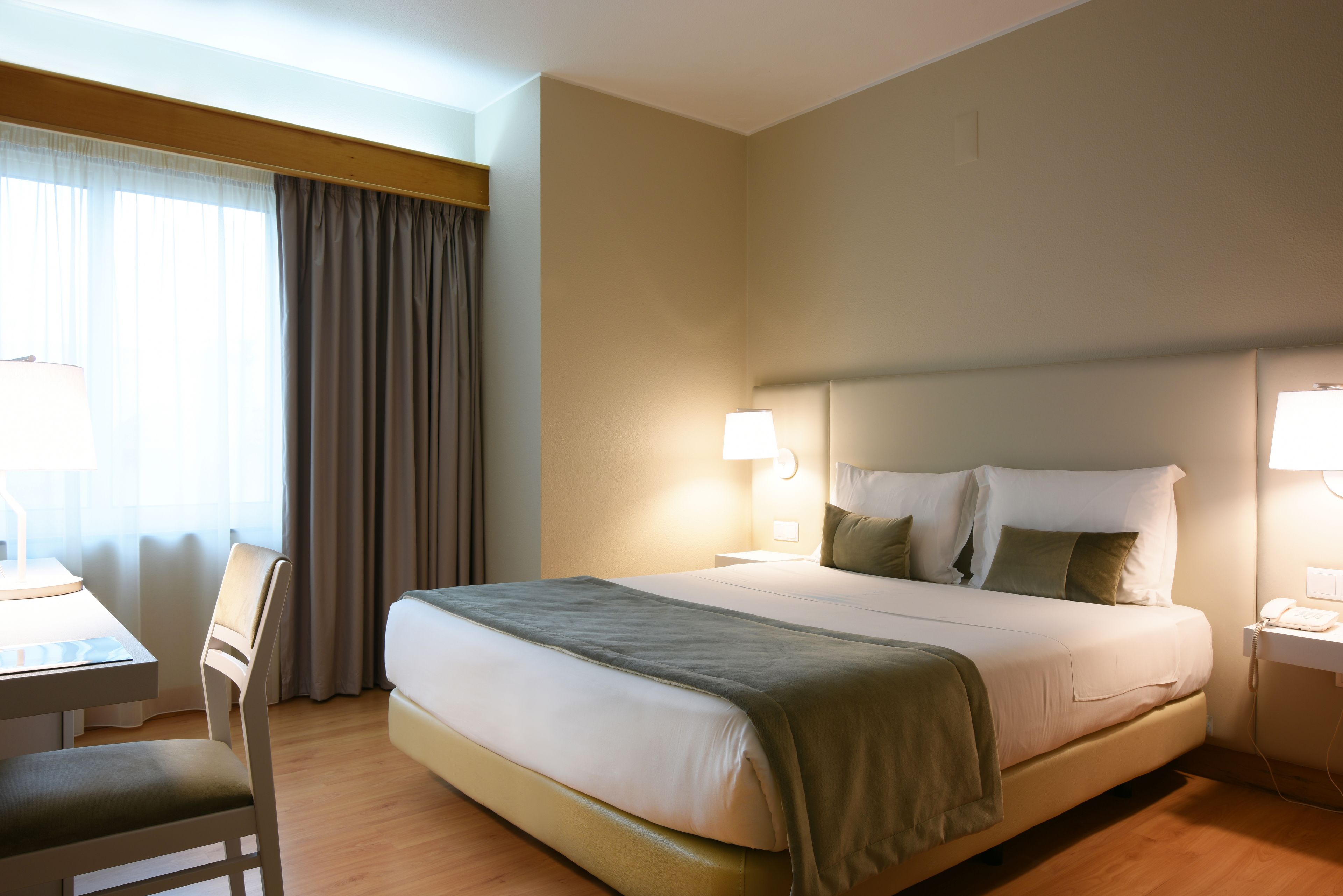 Une chambre d’hôtel comprenant un lit, des lampes de chevet, un bureau avec une chaise et une fenêtre avec des rideaux.