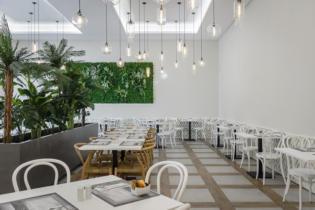A modern dining area with white tables and chairs, hanging pendant lights, and a green wall with plants.